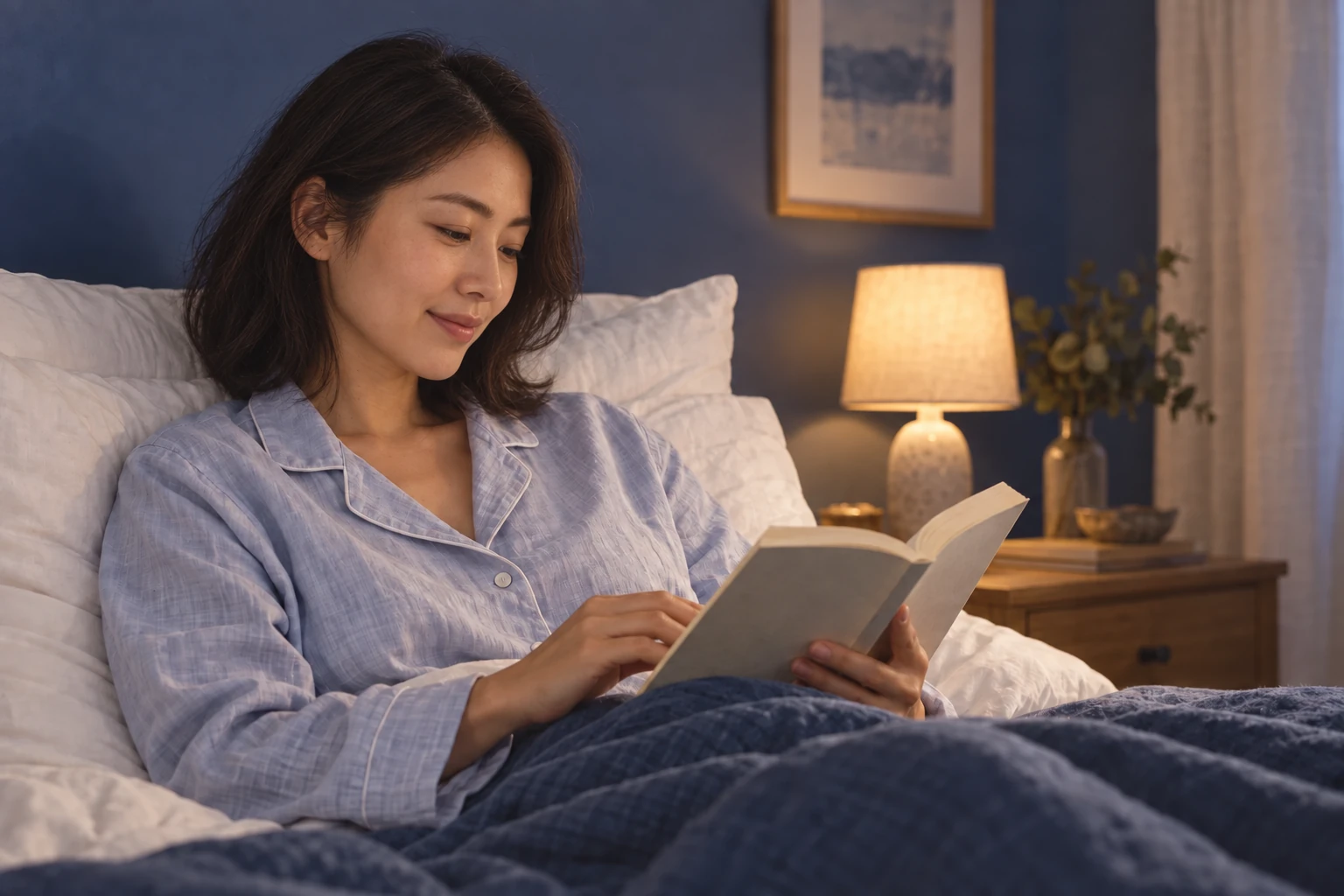 Illustrative portrait of a woman in her twenties reading in bed beside a softly lit lamp